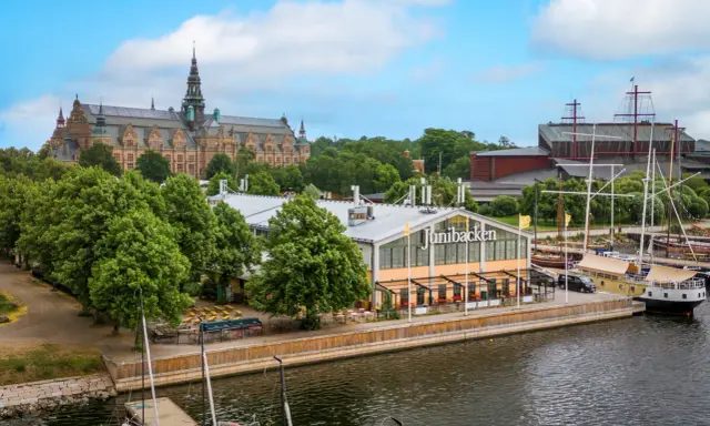 Junibacken, ett museum tillägnat Astrid Lindgren, ligger vid vattnet i Stockholm, med en stor byggnad i bakgrunden och segelfartyg i hamnen.