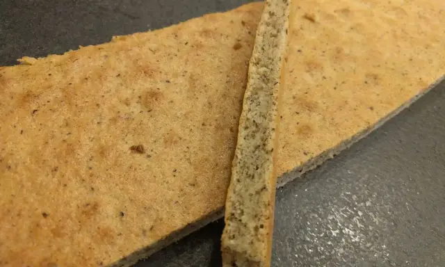 Deux fines tranches de biscuit joconde noisette, l'une posée sur l'autre, sur fond sombre.