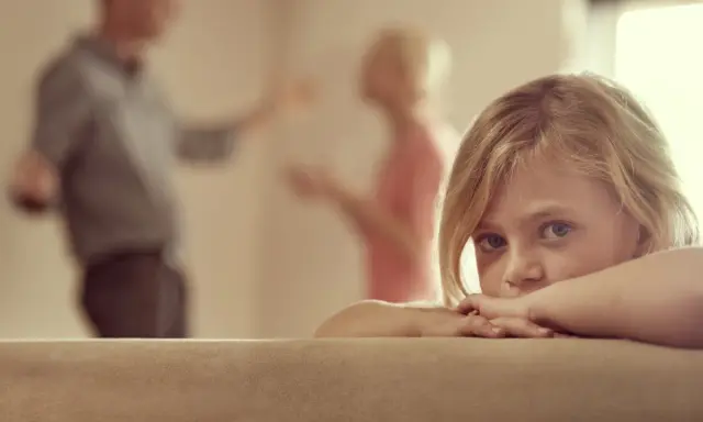 Niña asustada, escondida tras un sofá, mientras sus padres discuten delante de los hijos.