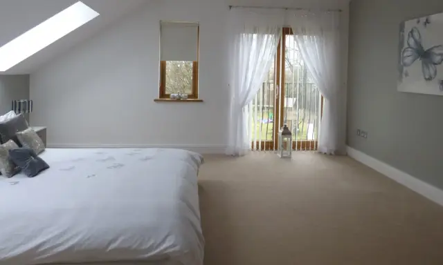 Dormitorio con cama blanca, ventana abuhardillada y puerta acristalada con cortinas. La orientación oeste de los pisos permite una luz suave.