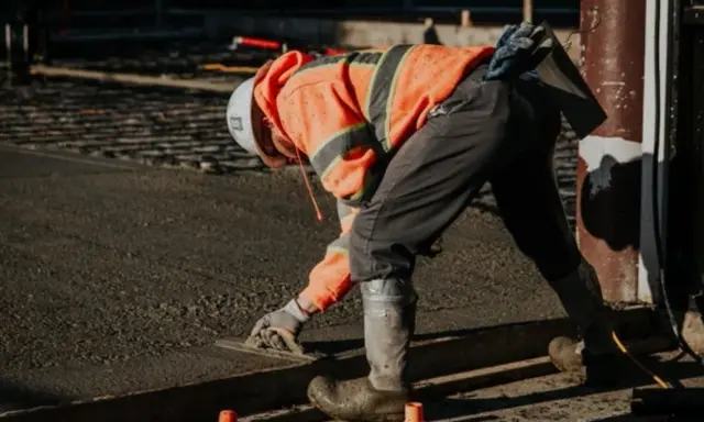Obrero con casco y chaleco naranja trabajando con hormigón autocompactante, alisando la superficie con una llana.