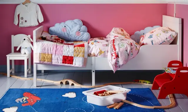 Habitación infantil con dos camas blancas, cojines de nube y mantas coloridas. Alfombra azul con tren de juguete y vías.
