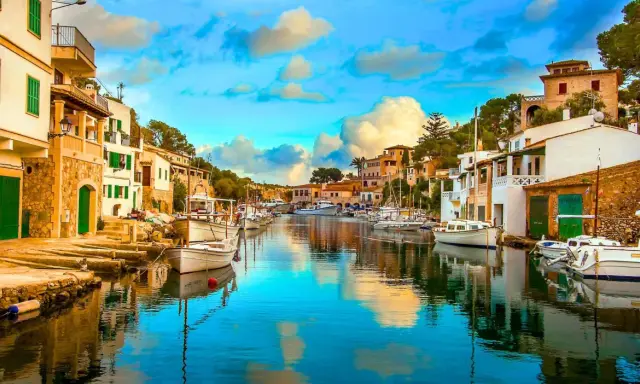 Un pintoresco pueblo pesquero con casas blancas y verdes, barcos amarrados en un canal de aguas cristalinas y un cielo azul con nubes esponjosas.