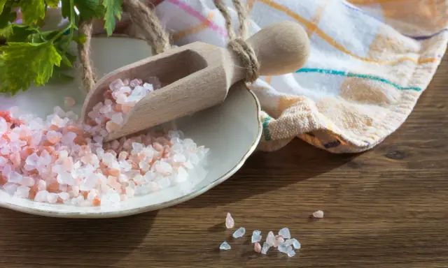Różowa sól himalajska w drewnianej łopatce na talerzyku, obok zielona gałązka i ręcznik.