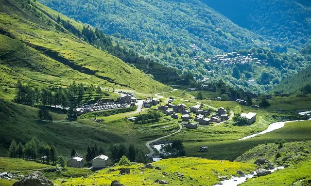 Un villaggio alpino nel **Pian della Regina**, con case in pietra e un fiume che scorre tra prati verdi e boschi lussureggianti.