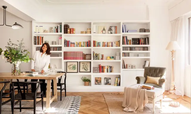 Una diseñadora casa sonríe junto a una mesa de comedor de madera, rodeada de estanterías llenas de libros y objetos decorativos.
