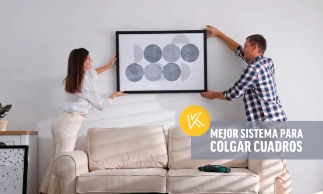 Pareja cuelga un cuadro moderno en pared de pladur. Un taladro descansa sobre el sofá, listo para la tarea.