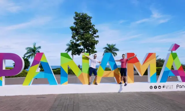 Two men pose with arms outstretched in front of a colorful "PANAMÁ" sign, suggesting Panama is gay friendly and a welcoming destination.