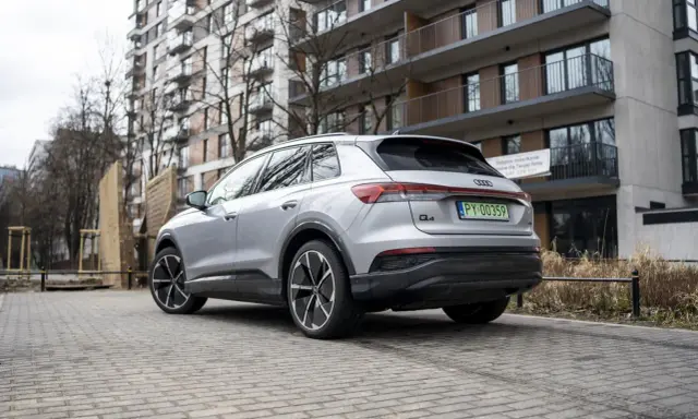 Srebrny Audi Q4 e-tron zaparkowany na brukowanym podjeździe przed nowoczesnym budynkiem mieszkalnym.