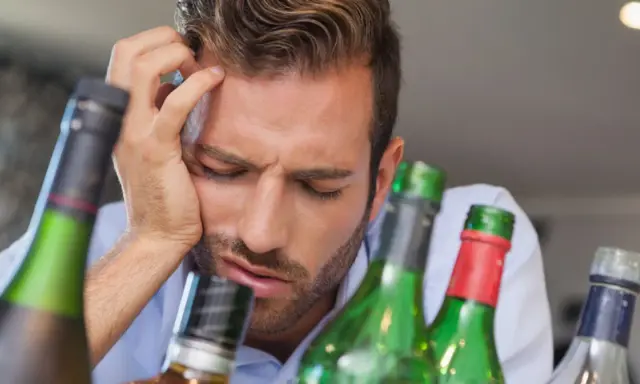 Mężczyzna z ręką na czole, otoczony butelkami, wyraźnie cierpi z powodu objawów przepicia alkoholowego.