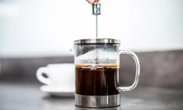 Eine Hand drückt den Stempel in eine French Press, um den perfekten Kaffee-Brew zuzubereiten. Eine Tasse wartet im Hintergrund.