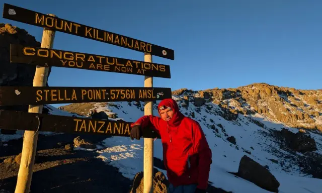 Mężczyzna w czerwonej kurtce przy znaku "Mount Kilimanjaro", najwyższy szczyt Afryki. Gratulacje, jesteś na Stella Point 5756 m n.p.m.