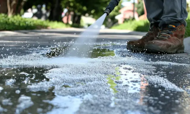 Dysza myjki ciśnieniowej spryskuje wodą i pianą chodnik, widać buty użytkownika.