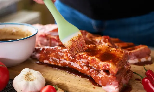 Pędzel rozprowadza aromatyczną marynatę do żeberek na surowym mięsie. Obok czosnek, pomidorki i papryczki chili.