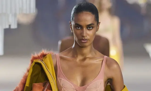 Modella in passerella con un audace reggiseno a vista color pesca, abbinato a una giacca gialla con pelliccia arancione.