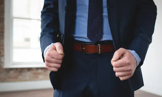 A man in a navy pinstripe suit and blue shirt wears a brown belt with black pants.