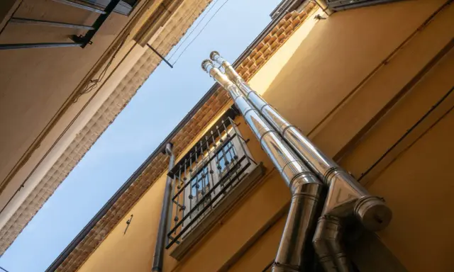 Dos tubos de salida humos estufa pellets brillan bajo el sol, ascendiendo por una pared amarilla junto a una ventana enrejada.