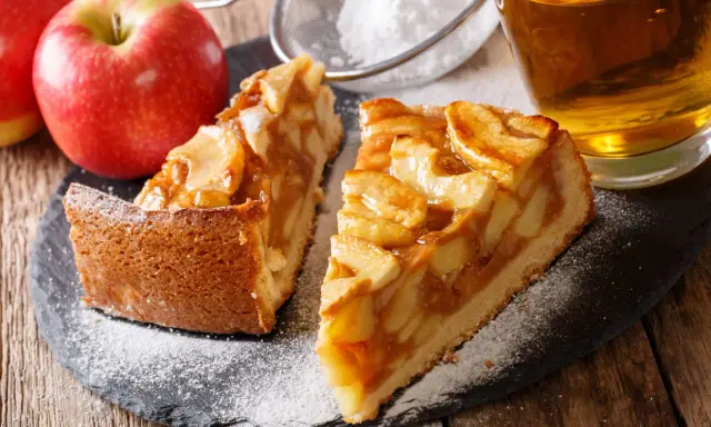 Deux parts de tarte aux pommes sur une ardoise, saupoudrées de sucre glace, avec des pommes et un verre de cidre.