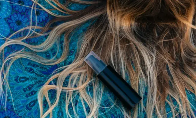 Czarny atomizer na tle błękitnego wzoru, obok rozpuszczonych, jasnych włosów. Idealny sposób, jak nawilżyć włosy.