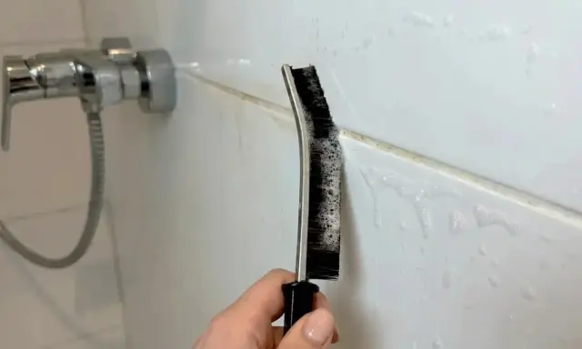 Hand reinigt Duschfugen mit einer Bürste und Schaum.