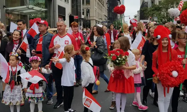 Dzieci i dorośli w strojach ludowych i z polskimi flagami maszerują ulicą. Widać serca z czerwonych balonów i kwiatów. Polacy w Szwecji świętują.