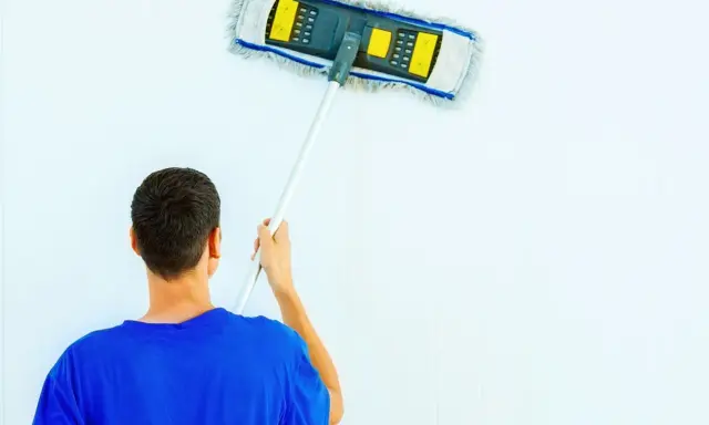 Hombre con camiseta azul limpiando paredes blancas con un plumero.