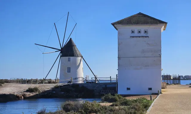 Altes Windrad und ein weißes Gebäude am Mar Menor, mit Wasser und Vegetation im Vordergrund.