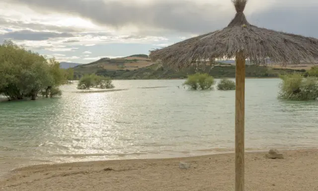 Sombrilla de paja en la orilla del pantano de Alloz. El agua turquesa, árboles emergiendo y colinas al fondo invitan a descubrir cómo llegar.