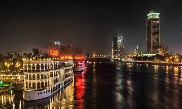 Beleuchtete Nilschiffe und moderne Hochhäuser spiegeln sich im Wasser. Ein magischer Anblick von Kairo bei Nacht.