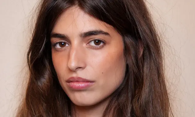 Portrait d'une jeune femme aux longs cheveux bruns. Son maquillage met en valeur ses yeux et ses lèvres, avec un soin particulier apporté à son sourcil poil à poil.