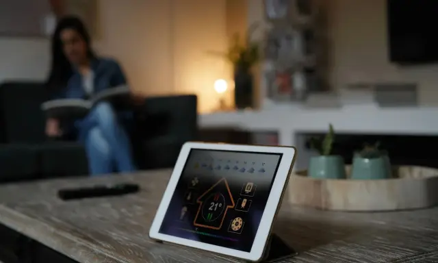 Tablette affichant l'interface pour installer un thermostat connecté. Une femme lit sur le canapé en arrière-plan.