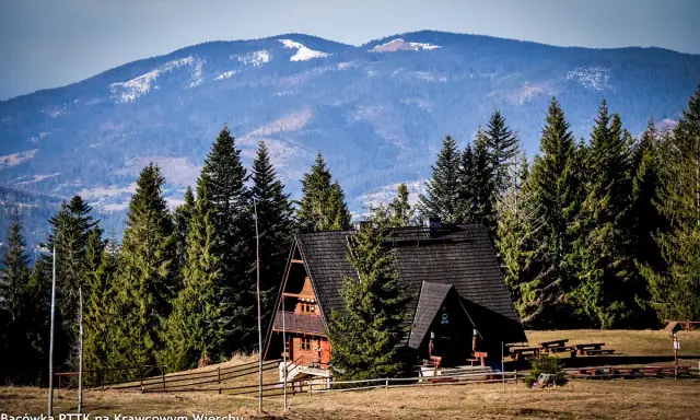 Drewniana bacówka PTTK na Krawcowym Wierchu, otoczona lasem świerkowym, z widokiem na ośnieżone góry.