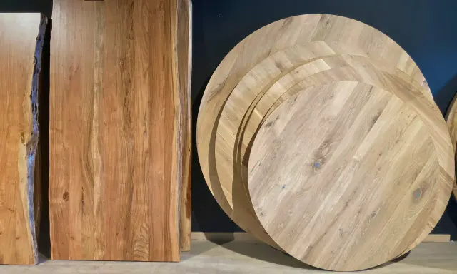 Plusieurs plateaux de table ronde en bois, empilés, illustrent la définition d'une table ronde. Des planches de bois brut sont aussi visibles.