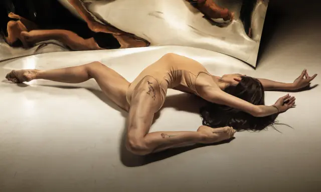 Młoda kobieta w cielistym body wygina się w łuk na podłodze, jej odbicie w lustrze tworzy surrealistyczny obraz. Jak z opowiadanie erotyczne nastolatki.
