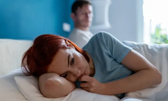 Zmartwiona kobieta leży w łóżku, podczas gdy jej partner jest niewyraźny w tle. Scena sugeruje **obsesyjne kontrolowanie partnera** i jego skutki.