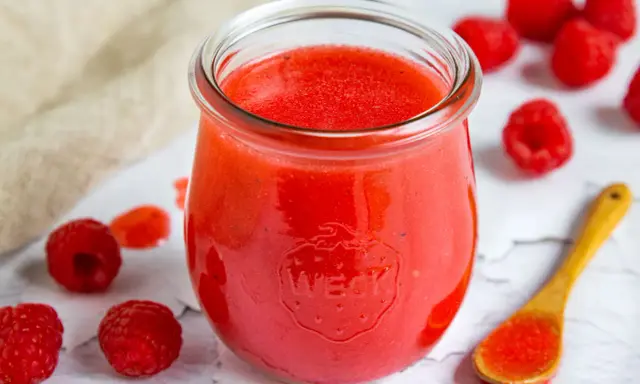 Ein Glas mit leuchtend roter Himbeer-Vinaigrette, umgeben von frischen Himbeeren und einem Holzlöffel mit etwas Vinaigrette.