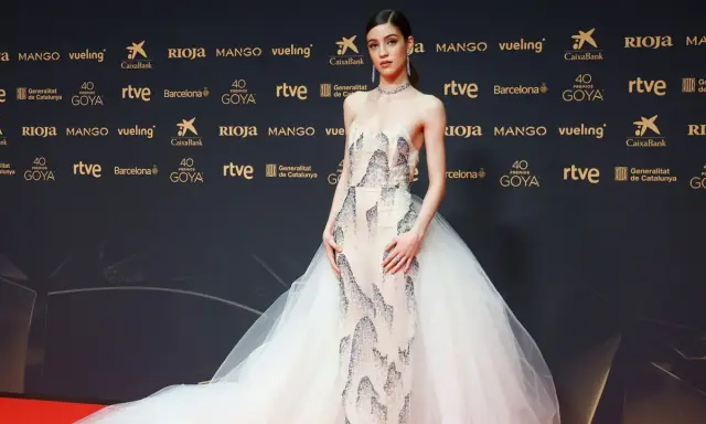 Una joven desfila en la goya alfombra roja con un vestido de gala blanco con detalles plateados y una voluminosa cola de tul.