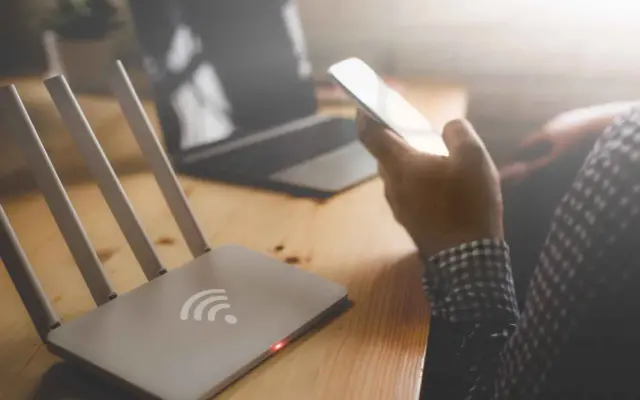 Router Wi-Fi, smartfon w dłoni, laptop na drewnianym biurku.