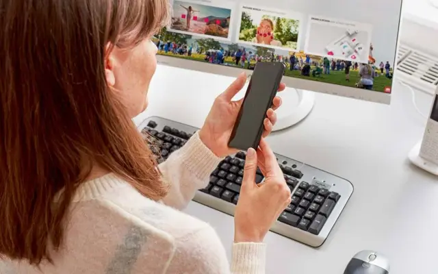 Kobieta trzyma smartfon przed monitorem komputera z klawiaturą i myszką.