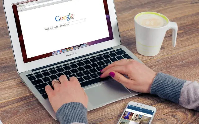 Kobieta pisze na laptopie z otwartą wyszukiwarką Google, obok stoi kubek kawy i smartfon.