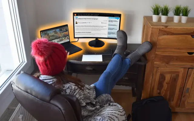Kobieta w czerwonej czapce i szarych kozakach pracuje przy laptopie i zewnętrznym monitorze.