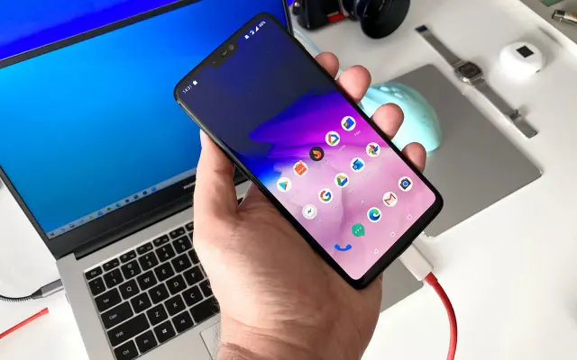 Dłoń trzyma smartfon podłączony kablem do laptopa.