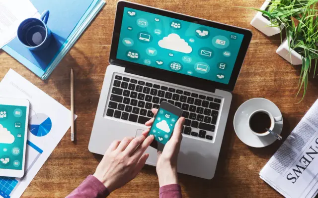 Dłonie trzymające smartfon z ikonami chmury na tle laptopa i dokumentów.
