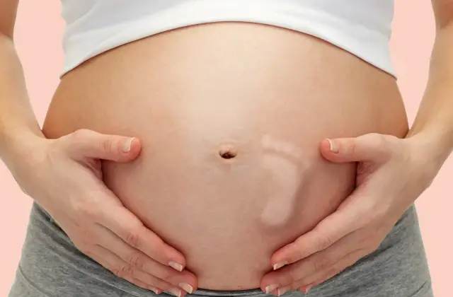 Brzuszek w 29 tygodniu ciąży, z widocznym odciskiem stópki dziecka. Ręce delikatnie obejmują brzuch.