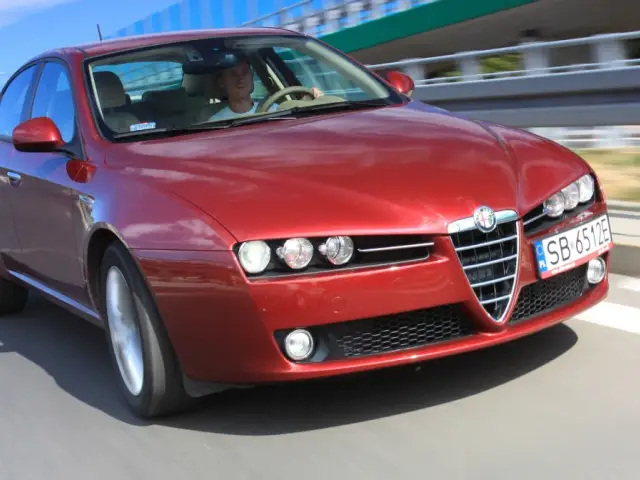 Czerwone Alfa Romeo 159 1.9 JTD pędzi po drodze, kierowca skupiony na trasie.