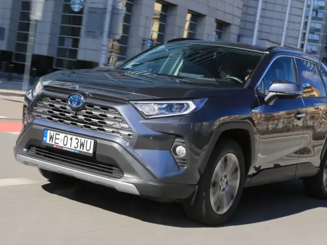 Ciemnoszary Toyota RAV4 Hybrid pędzi po mieście, odbijając w lustrach budynki i niebo.