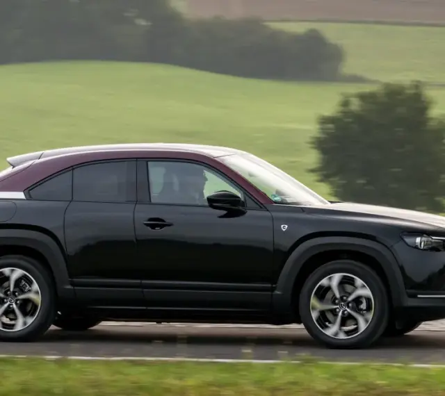 Czarna Mazda MX-30 z bordowym dachem sunie po drodze wśród zielonych wzgórz.
