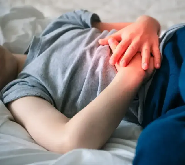 Dziecko leży w łóżku, trzymając się za brzuch. Widać zaczerwienienie sugerujące ból brzucha u dziecka.