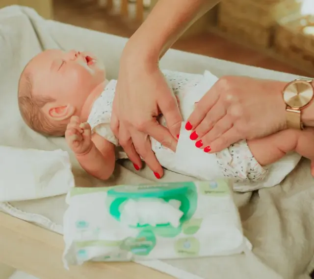 Delikatne dłonie z czerwonymi paznokciami pokazują, jak przewijać noworodka, używając chusteczek nawilżanych.