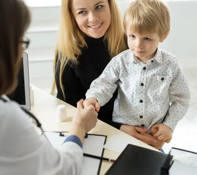 Mama z synkiem u lekarza. Chłopiec ściska dłoń lekarce, a mama uśmiecha się. W tle widać tablet i białą buteleczkę, jakby z lekiem. To ważny moment, być może wpis do książeczki zdrowia.
